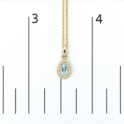 Close-up image of a yellow metal necklace pendant with a pear-shaped blue gemstone surrounded by white round gemstones in a prong setting, shown on a scale.