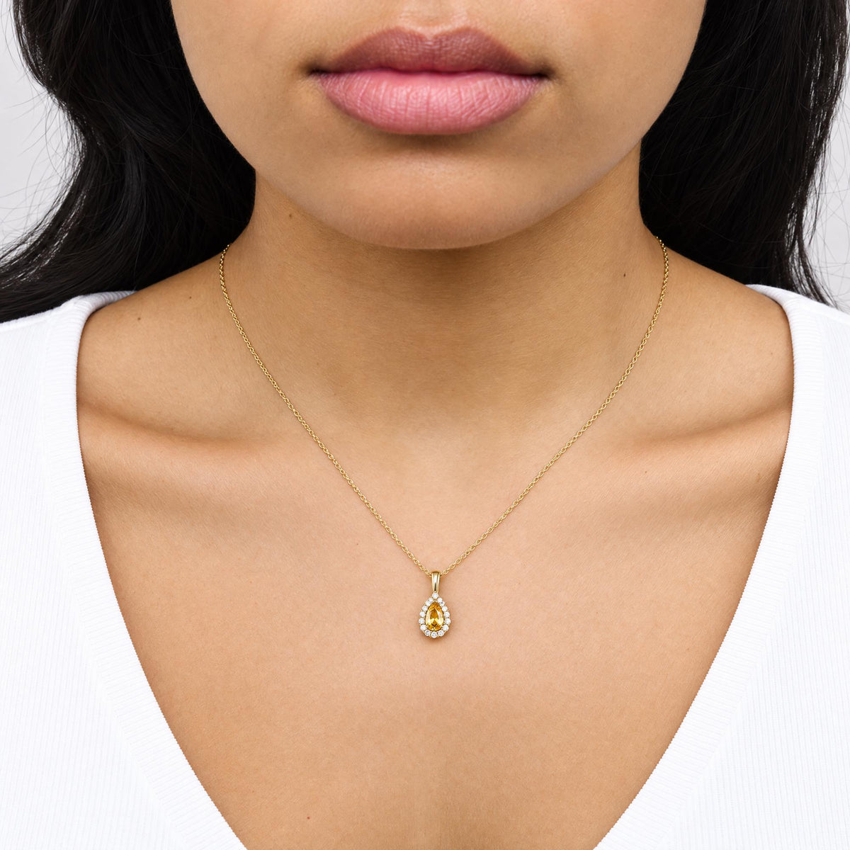 Close-up front view of a model wearing a yellow metal necklace featuring a pear-shaped yellow gemstone pendant surrounded by round white gemstones in a prong setting.