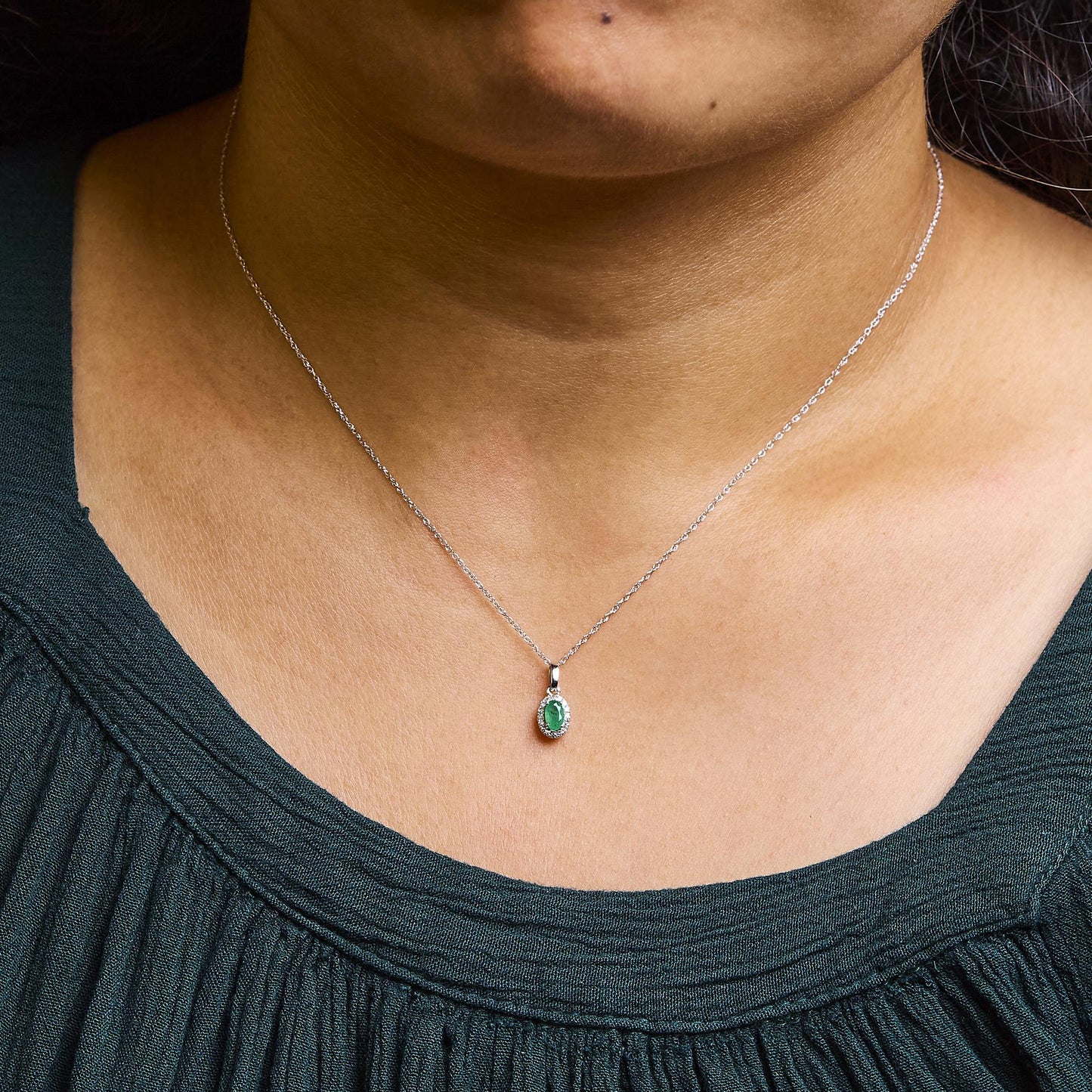 White metal necklace with oval green gemstone pendant surrounded by small round white gemstones in micro-pave setting, shown on a model from the front.