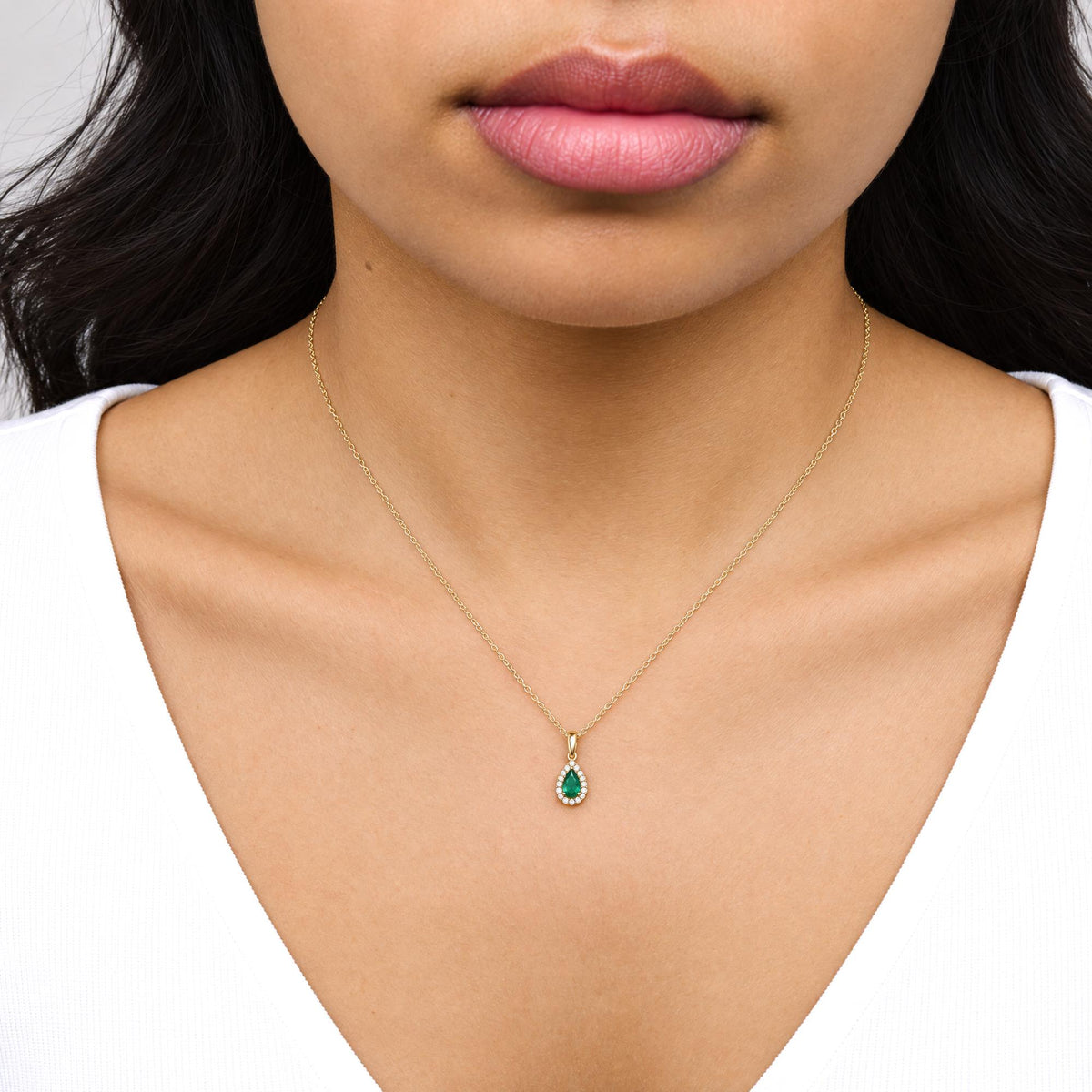 Close-up front view of a model wearing a yellow metal necklace with a pear-shaped green gemstone pendant surrounded by small round white gemstones.
