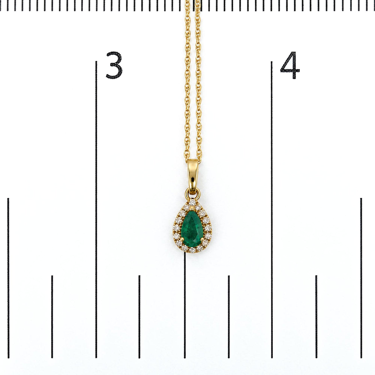 Close-up front view of a yellow metal necklace pendant featuring a pear-shaped green gemstone surrounded by round white gemstones in a prong setting, shown on a scale.