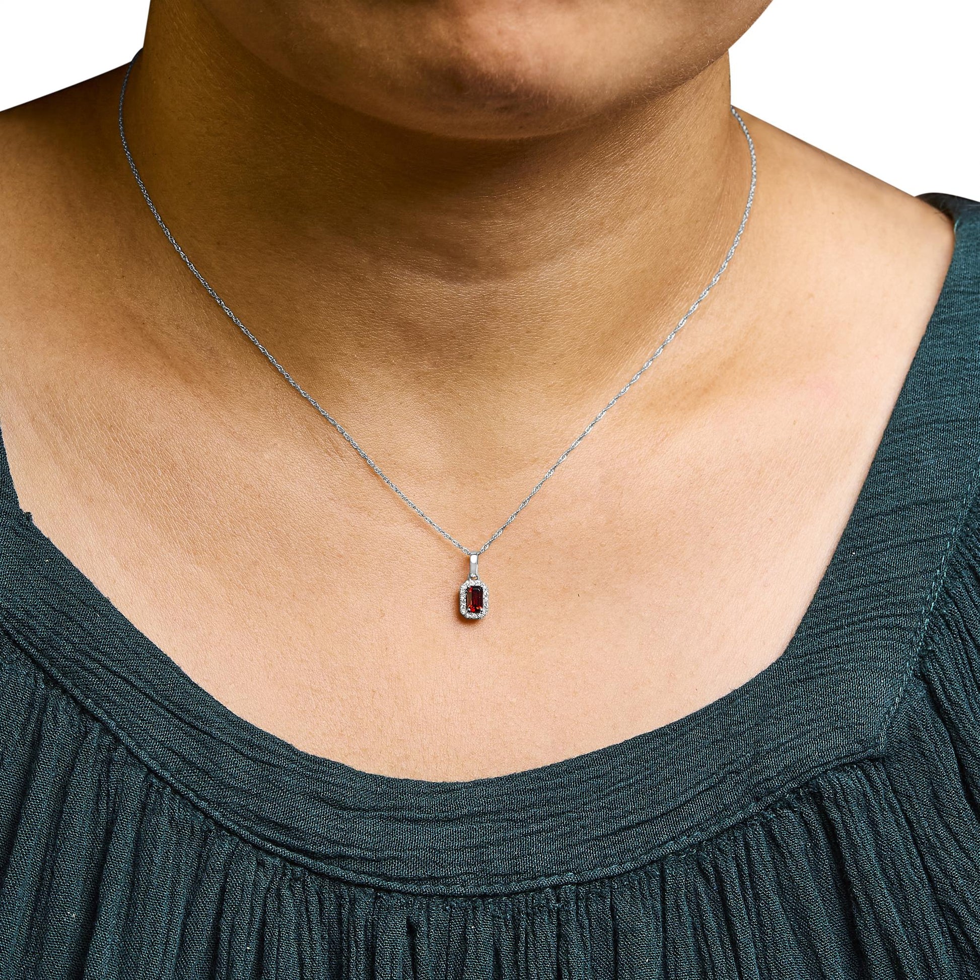 Close-up front view of a white metal necklace on a model, featuring a pendant with a red gemstone in an emerald shape, surrounded by small round white gemstones in micro-pave setting.