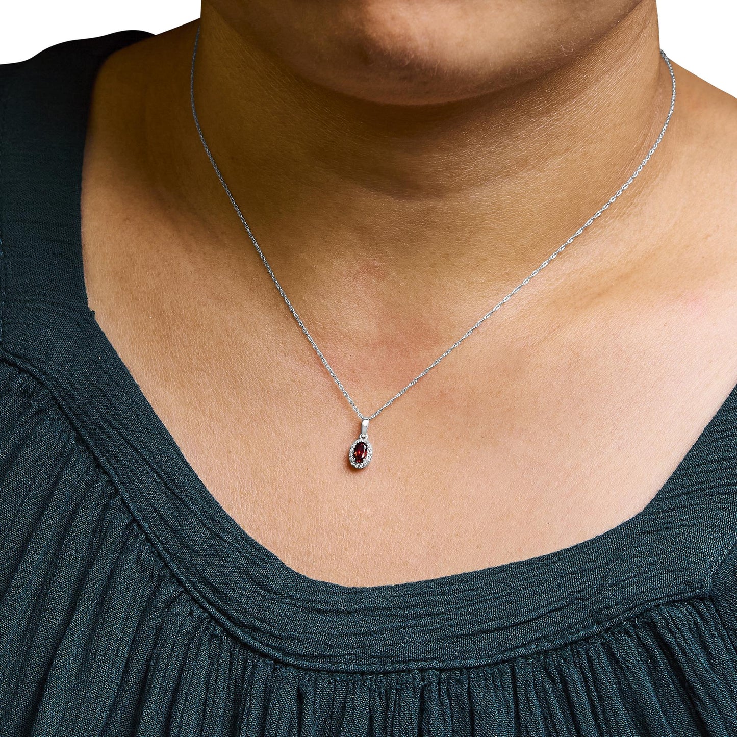 Close-up front view of a white metal necklace on a model featuring a small oval red gemstone pendant surrounded by small round white gemstones in a micro-pave setting.