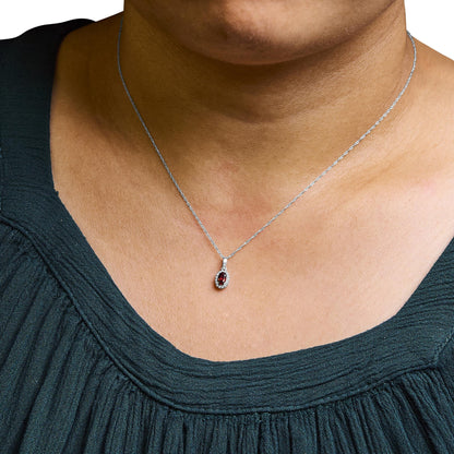Close-up front view of a white metal necklace on a model featuring a small oval red gemstone pendant surrounded by small round white gemstones in a micro-pave setting.