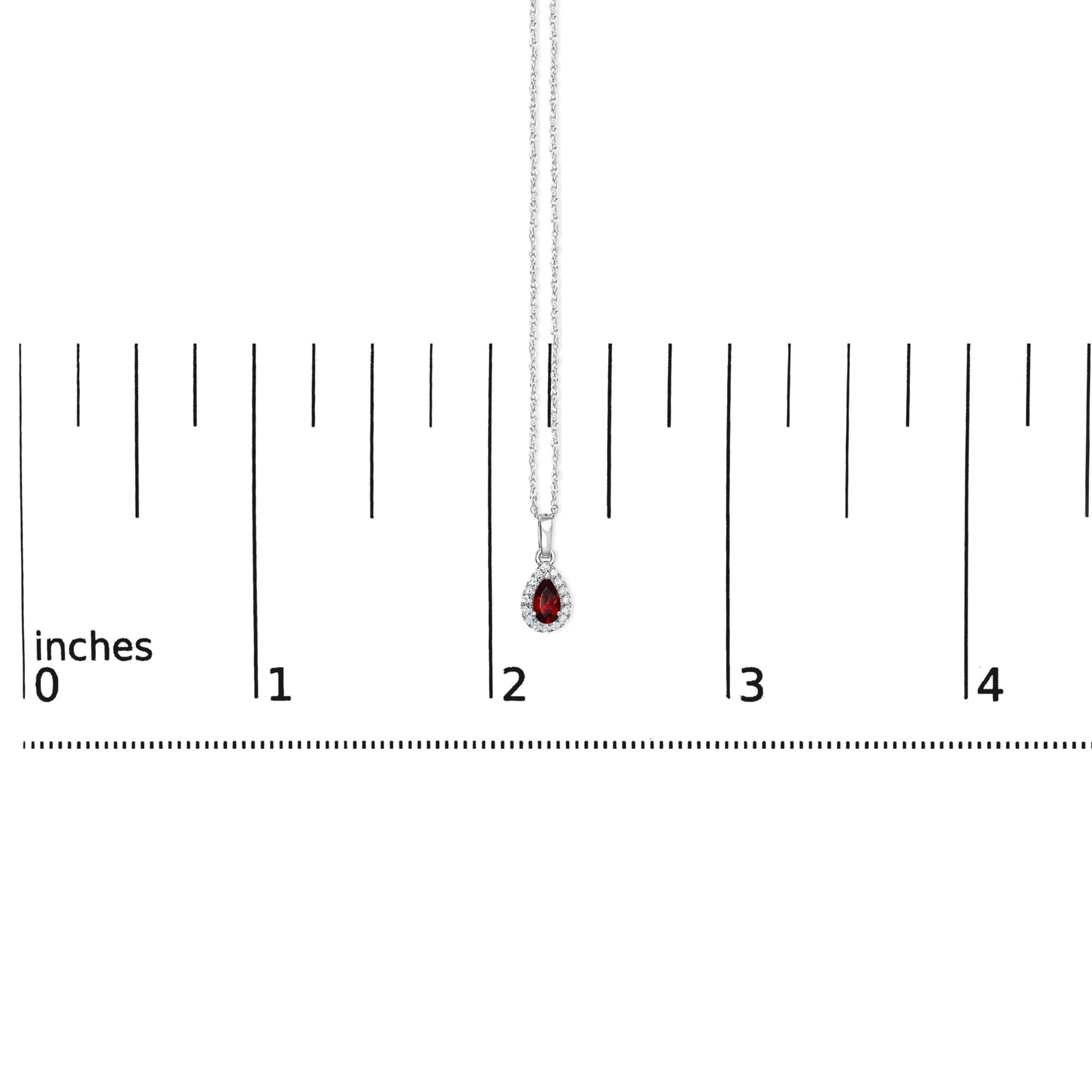 White metal necklace with a pear-shaped red gemstone pendant surrounded by round white gemstones, shown hanging vertically over a ruler for size reference.