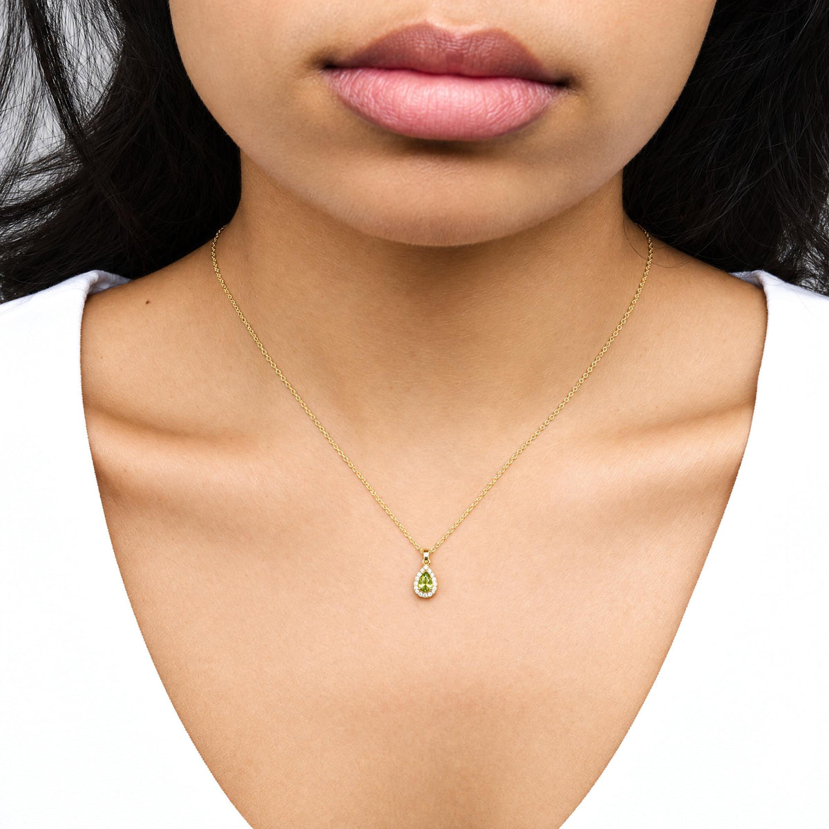 Close-up front view of a model wearing a yellow metal necklace with a pear-shaped green gemstone pendant surrounded by round white gemstones in prong settings.