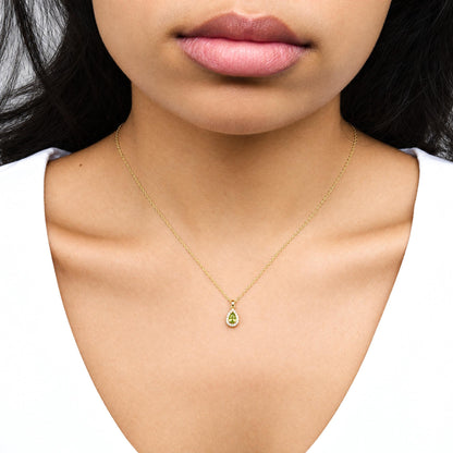 Close-up front view of a model wearing a yellow metal necklace with a pear-shaped green gemstone pendant surrounded by round white gemstones in prong settings.