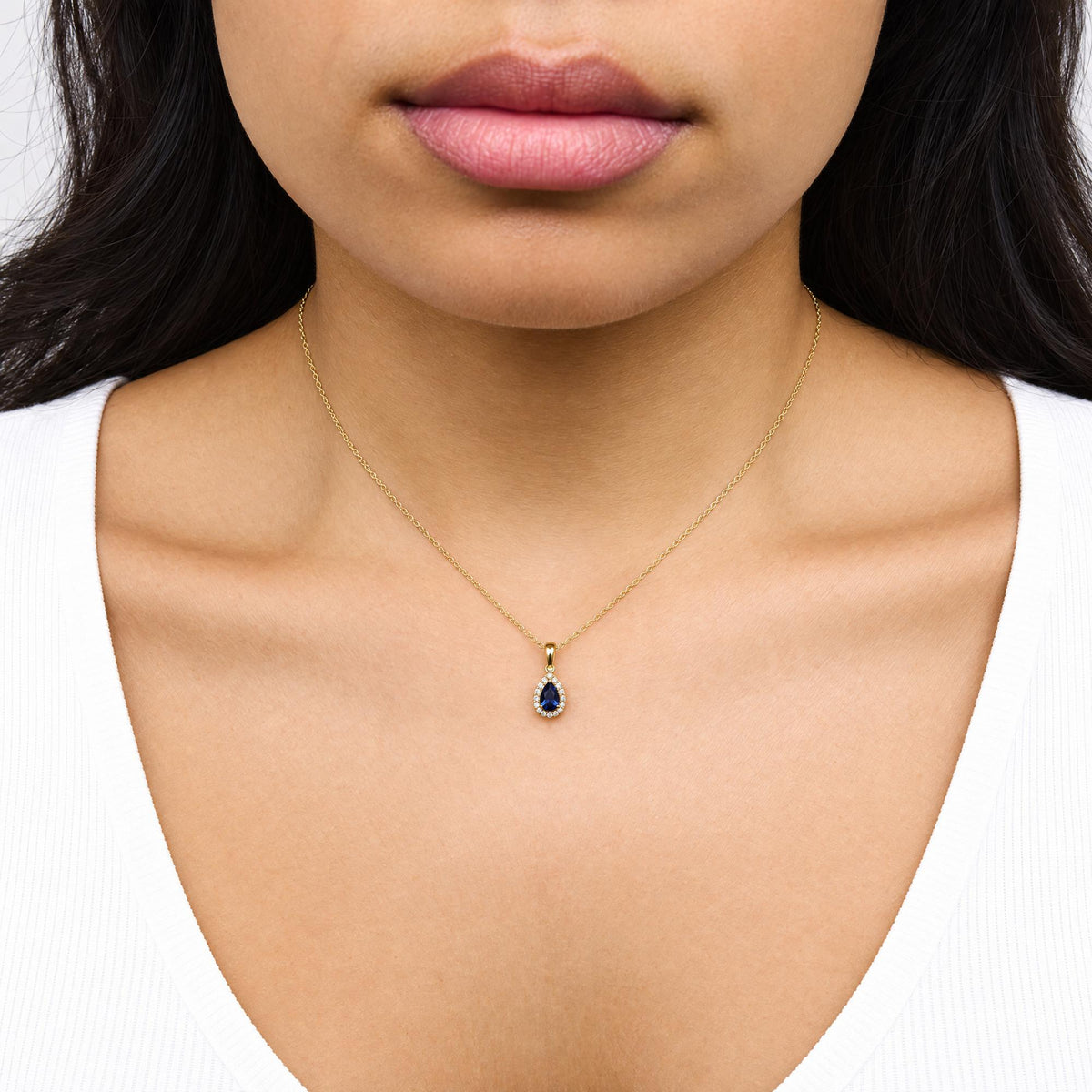 Close-up front view of a model wearing a yellow metal necklace with a pear-shaped blue gemstone pendant surrounded by small white gemstones.