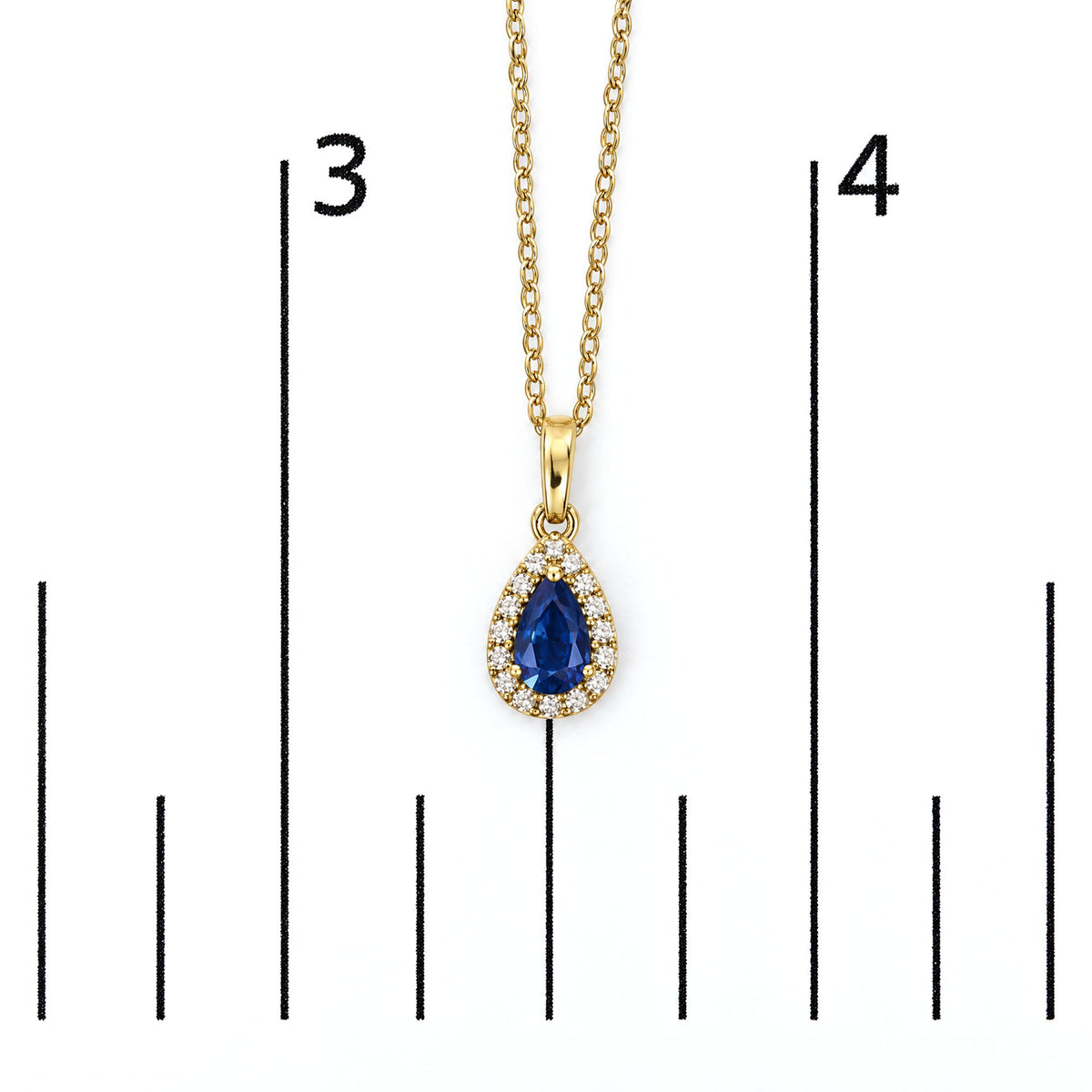 Close-up front view of a yellow metal necklace pendant with a pear-shaped blue gemstone surrounded by small round white gemstones, shown on a scale.
