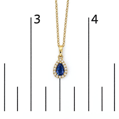 Close-up front view of a yellow metal necklace pendant with a pear-shaped blue gemstone surrounded by small round white gemstones, shown on a scale.