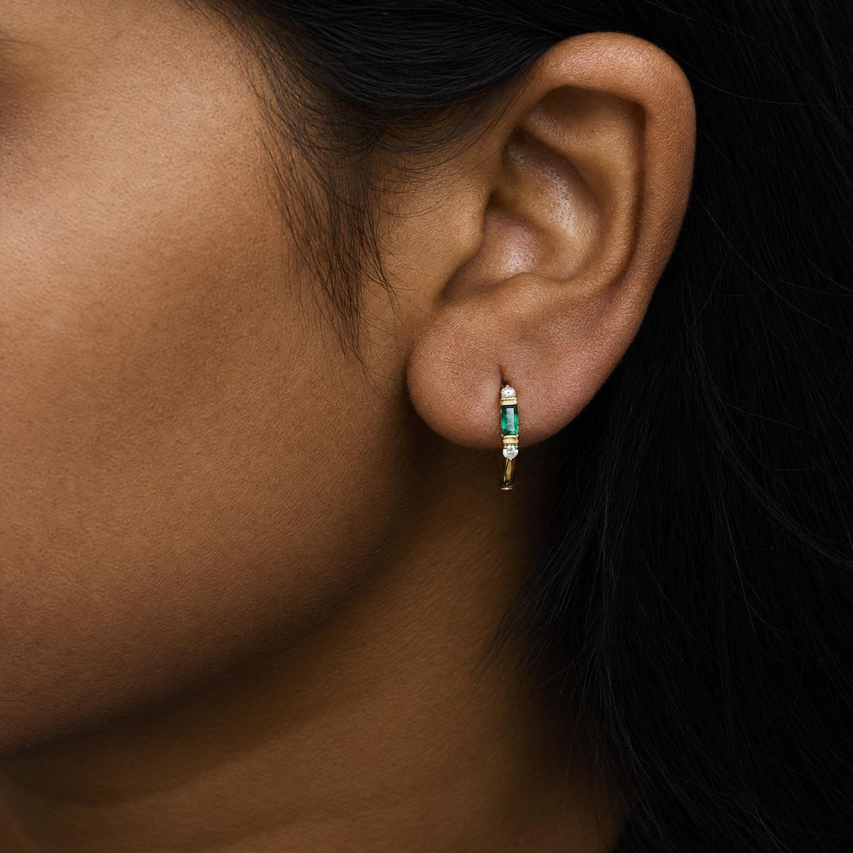 Close-up side view of a model wearing yellow metal earrings featuring a vertical green baguette-shaped gemstone in channel setting, flanked by round white gemstones in prong setting.