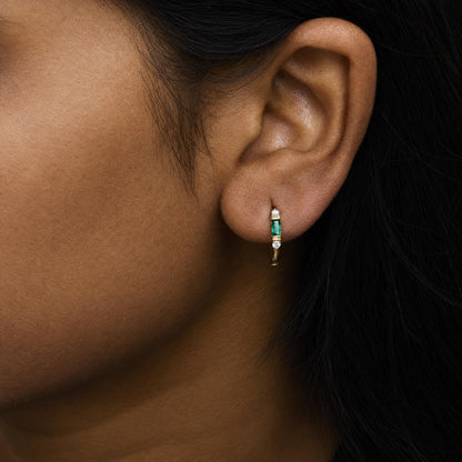 Close-up side view of a model wearing yellow metal earrings featuring a vertical green baguette-shaped gemstone in channel setting, flanked by round white gemstones in prong setting.