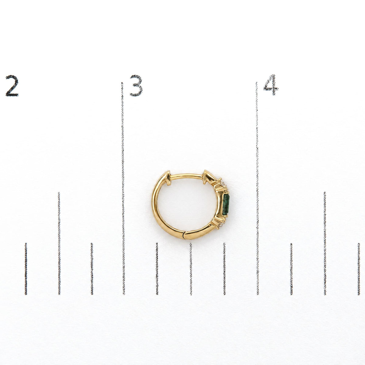 Close-up top view of a single yellow hoop earring shown on a scale, featuring round white gemstones in prong settings and green baguette-shaped gemstones in channel settings.