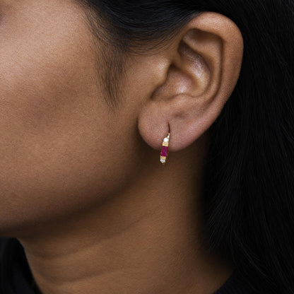 Close-up side view of a model wearing a yellow metal hoop earring featuring round white gemstones in prong settings and baguette red gemstones in channel settings.