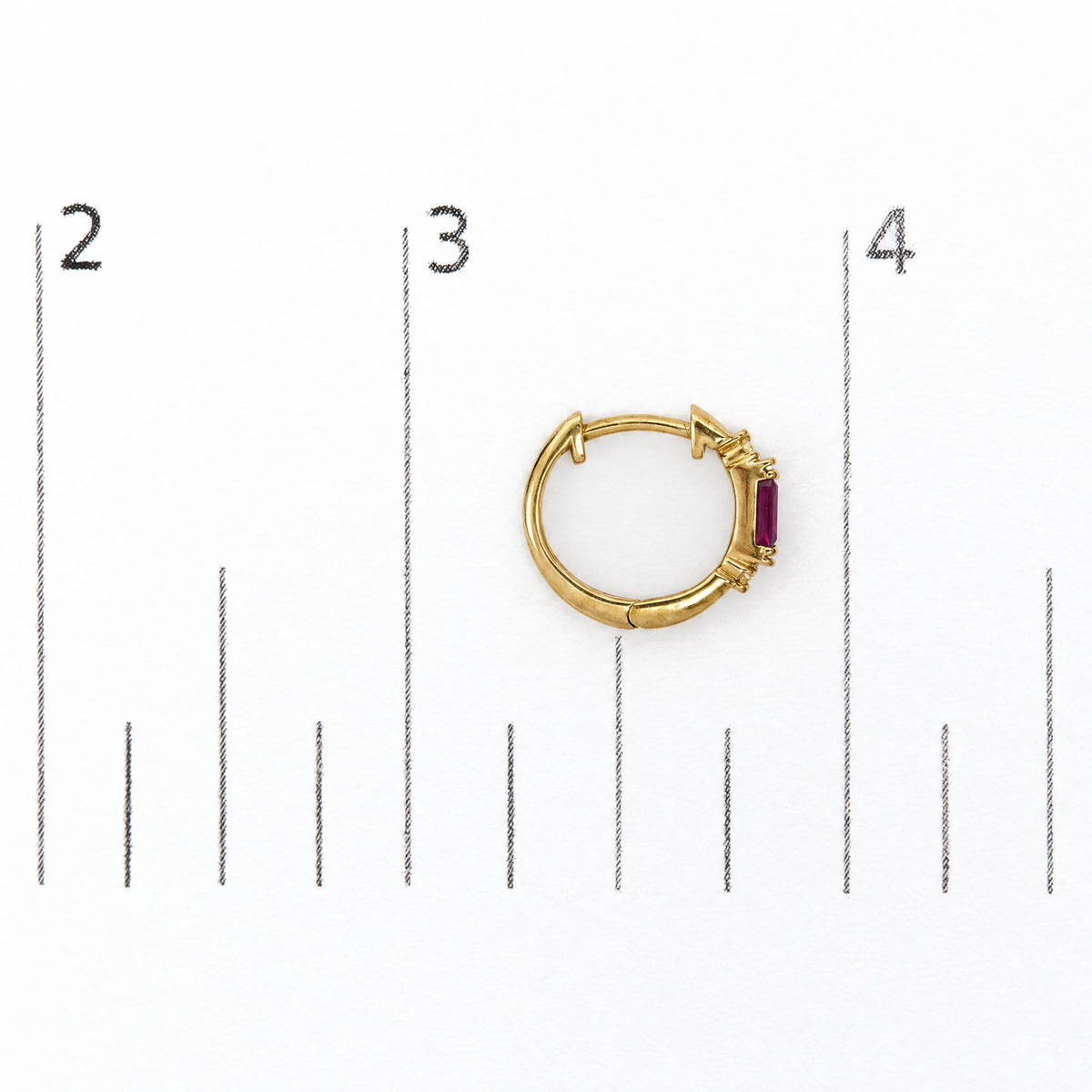Close-up top view of a yellow metal hoop earring set with a red baguette-shaped gemstone in a channel setting and four round white gemstones in prong settings, shown on a white background with a measurement scale.