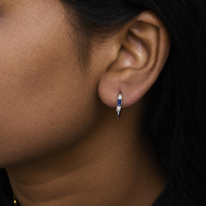 Close-up side view of a model wearing white earrings featuring two blue baguette-shaped gemstones in a channel setting and four round white gemstones in prong settings.