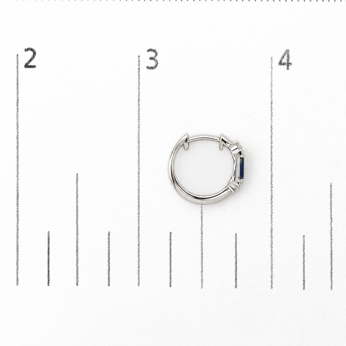 Close-up top view of a white metal hoop earring shown on a measurement scale, featuring round white gemstones in prong settings and blue baguette-shaped gemstones in a channel setting.