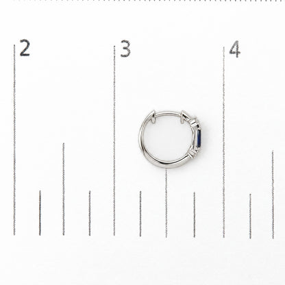 Close-up top view of a white metal hoop earring shown on a measurement scale, featuring round white gemstones in prong settings and blue baguette-shaped gemstones in a channel setting.