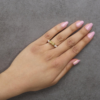Yellow ring with textured design and small white gemstones, worn on a model's finger shown from above on a gray textured background.