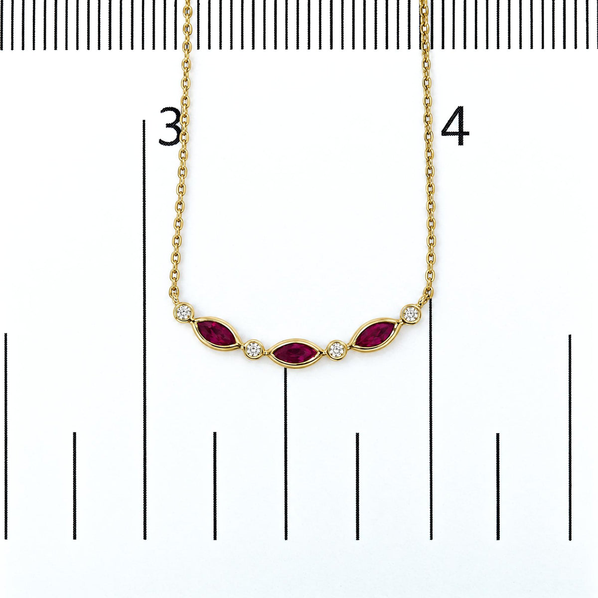 Close-up overhead shot of a yellow metal necklace with three marquise-shaped red gemstones in prong settings, interspersed with round white gemstones in bezel settings, shown on a scale.