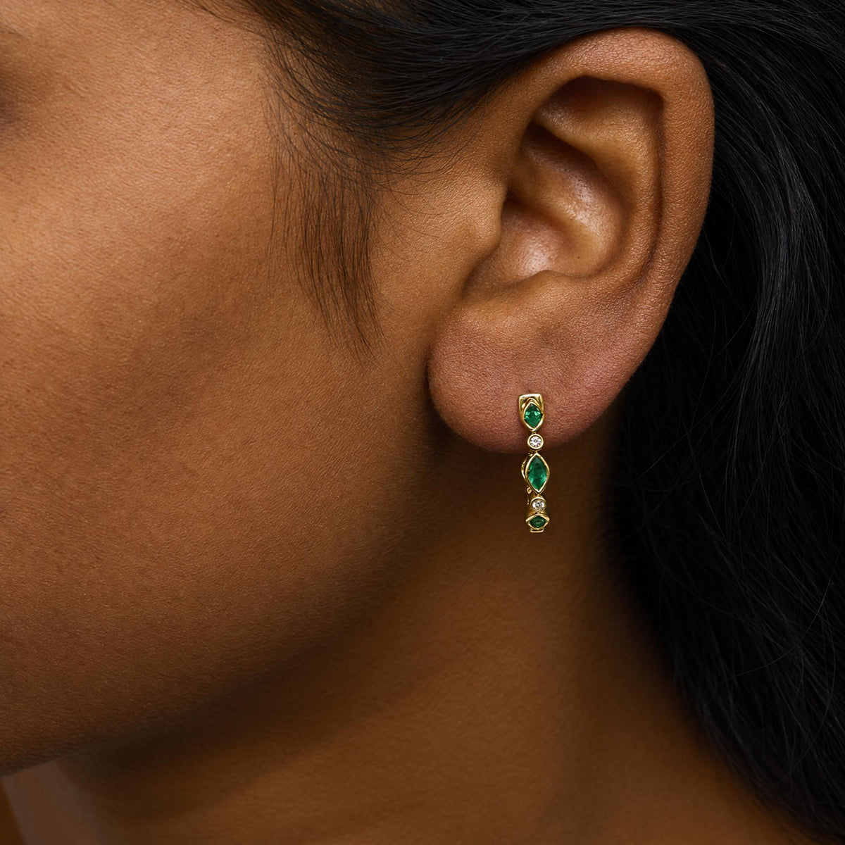 Close-up side view of a model wearing a yellow metal earring featuring bezel-set green marquise-shaped gemstones and white round gemstones arranged vertically.