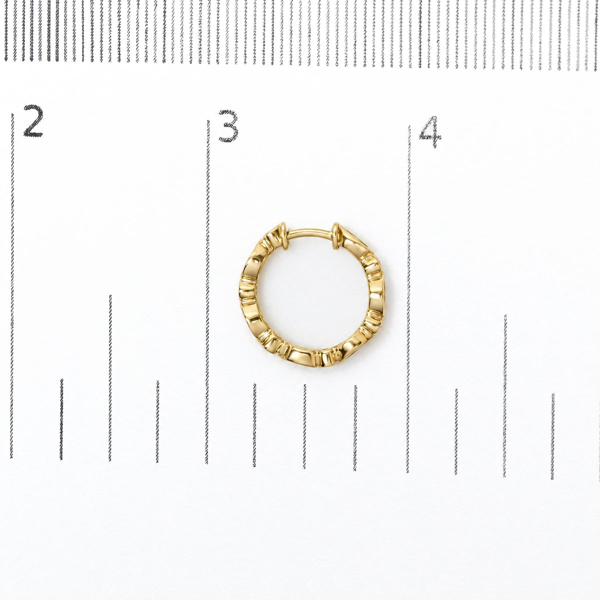 Close-up front view of a single yellow metal hoop earring with textured ridges, placed on a measurement scale.