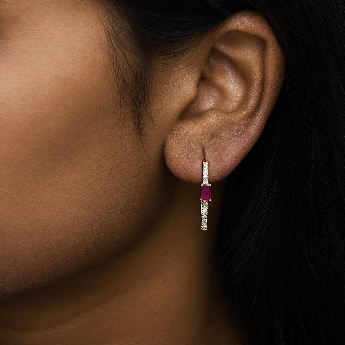 Close-up angled view of a model's ear wearing a yellow metal earring featuring a vertical row of small white round gemstones and a single larger red gemstone in prong settings.