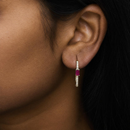 Close-up angled view of a model's ear wearing a yellow metal earring featuring a vertical row of small white round gemstones and a single larger red gemstone in prong settings.