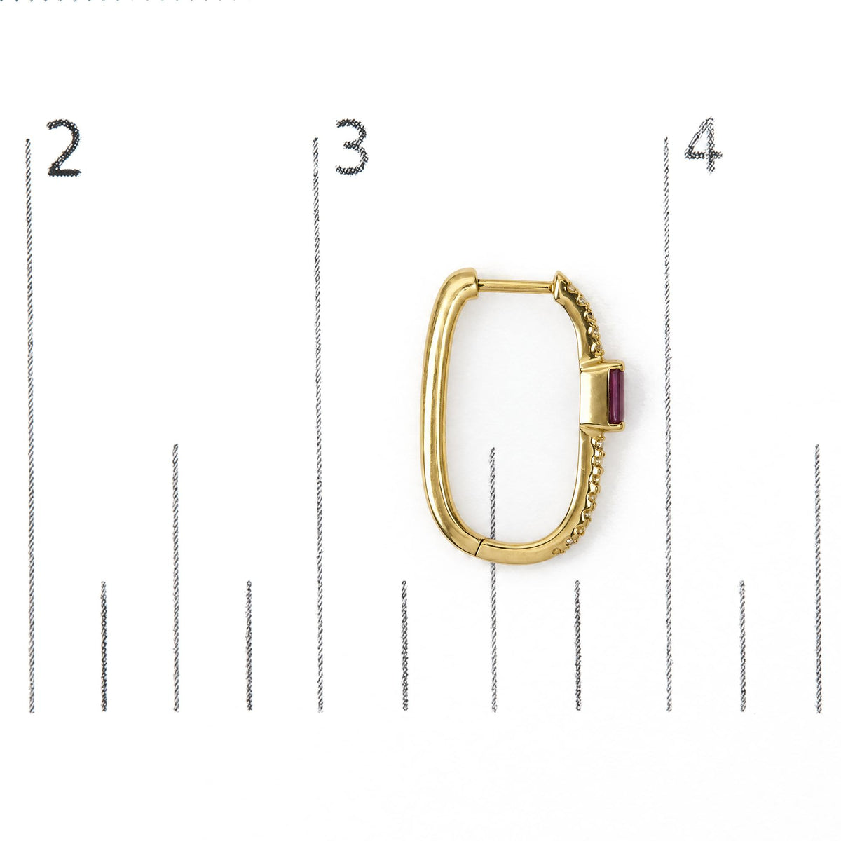 Close-up front angle of a single yellow metal earring shown on a scale, oval-shaped hoop design with small round white gemstones in prong settings along one side and a red rectangular gemstone set in prongs near the clasp.