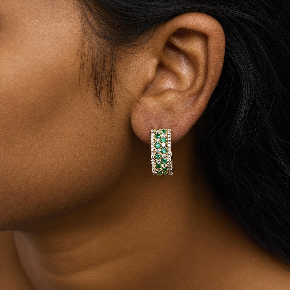 Close-up side view of a model wearing a yellow metal earring featuring a wide rectangular shape with multiple rows of round green and white gemstones in prong settings.