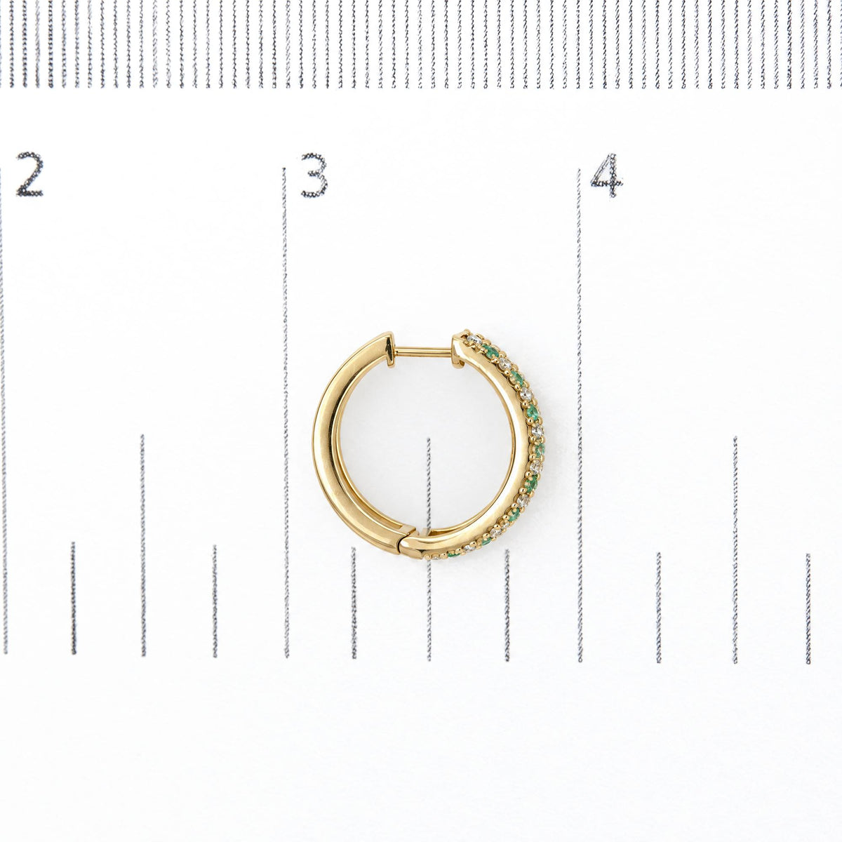 Close-up front view of a yellow metal hoop earring shown on a ruler scale, featuring alternating round white and green gemstones in prong settings along the outer edge.