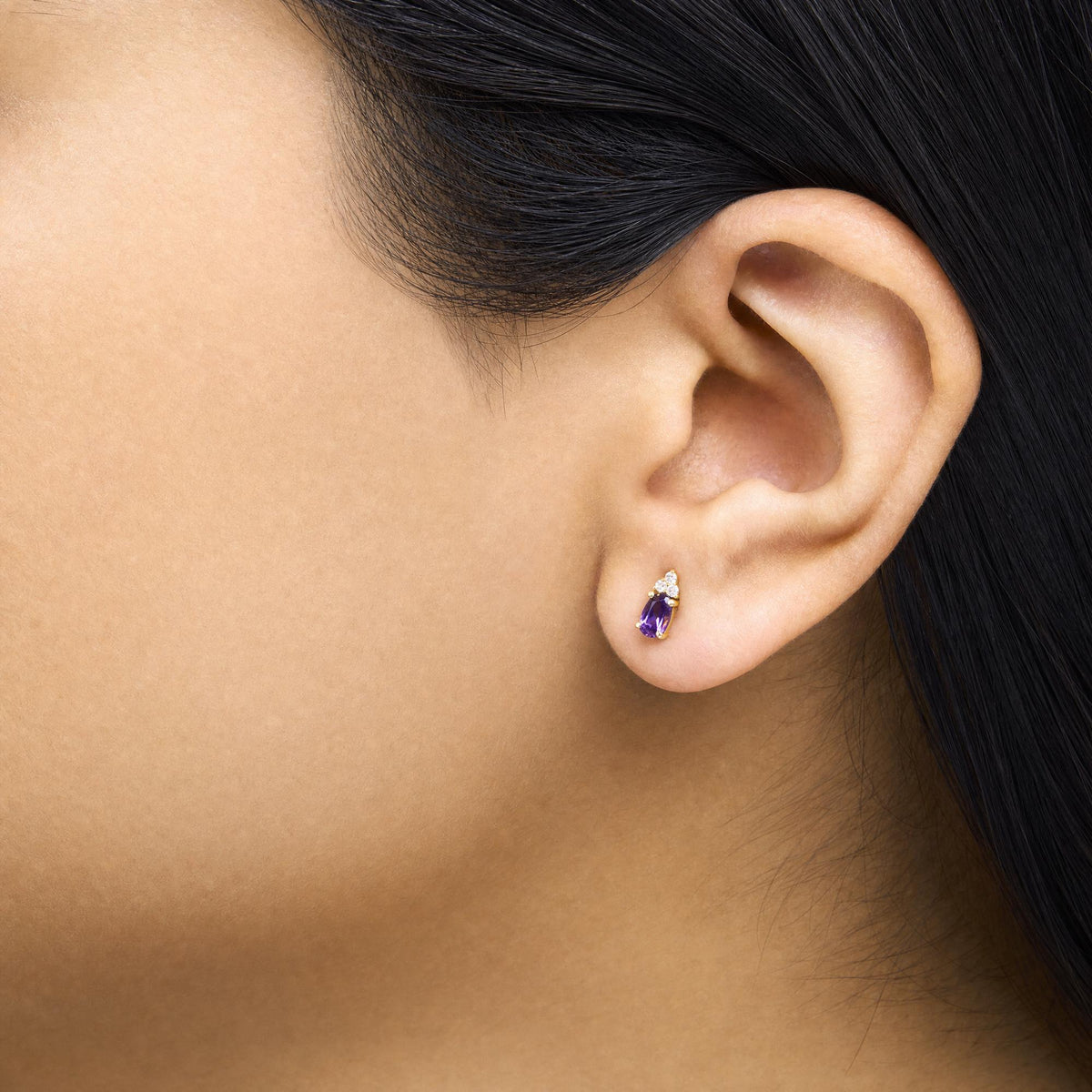Close-up side view of a model wearing yellow metal earrings featuring an oval purple gemstone and round white gemstones in a prong setting.