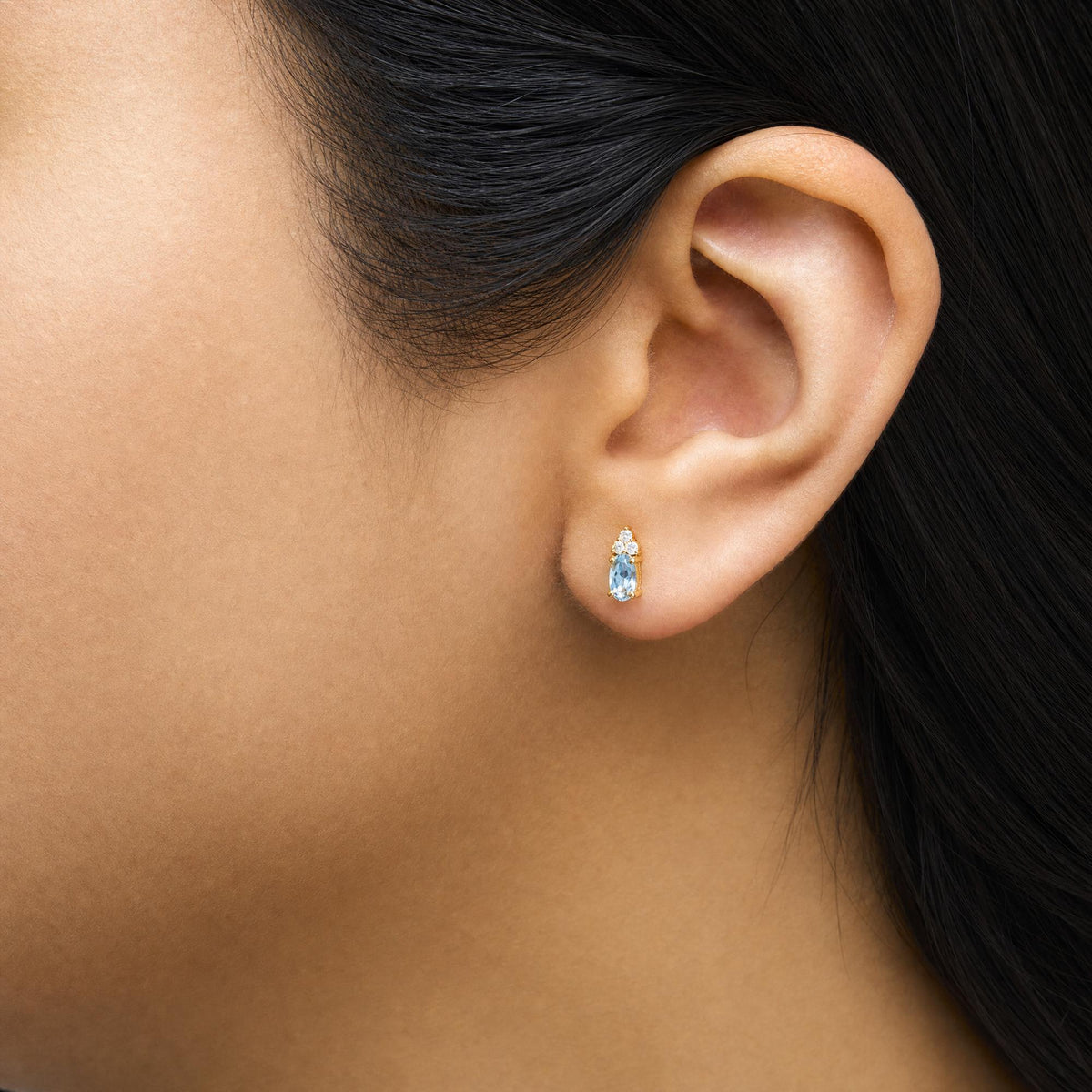 Close-up side view of a model wearing yellow metal earrings featuring a blue oval gemstone and multiple round white gemstones arranged above it in a prong setting.