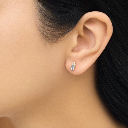 Close-up side view of a model's ear wearing yellow metal earrings featuring an oval blue gemstone and multiple small round white gemstones in prong settings.