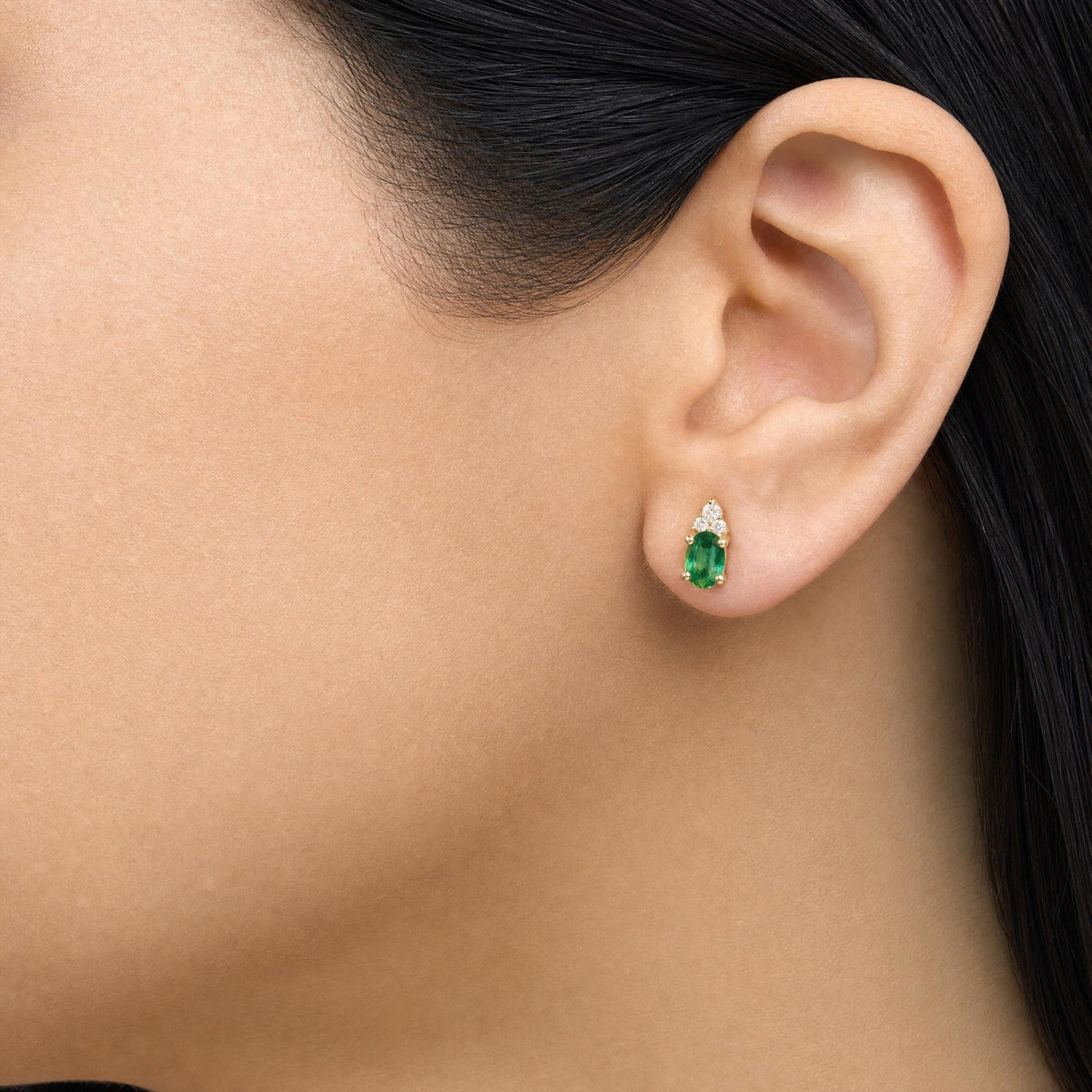 Close-up side view of a model wearing yellow metal earrings featuring an oval green gemstone at the bottom and round white gemstones arranged above it in a prong setting.