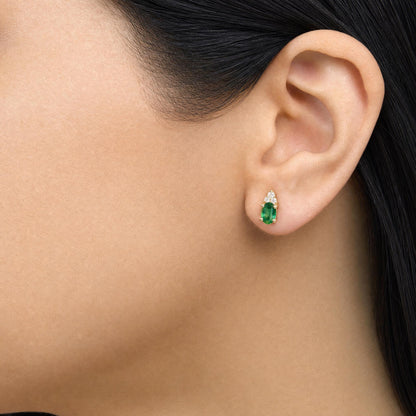 Close-up side view of a model wearing yellow metal earrings featuring an oval green gemstone at the bottom and round white gemstones arranged above it in a prong setting.