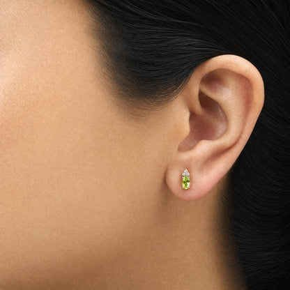 Close-up side view of a model wearing yellow metal earrings featuring an oval green gemstone with prong setting and a cluster of round white gemstones above it.