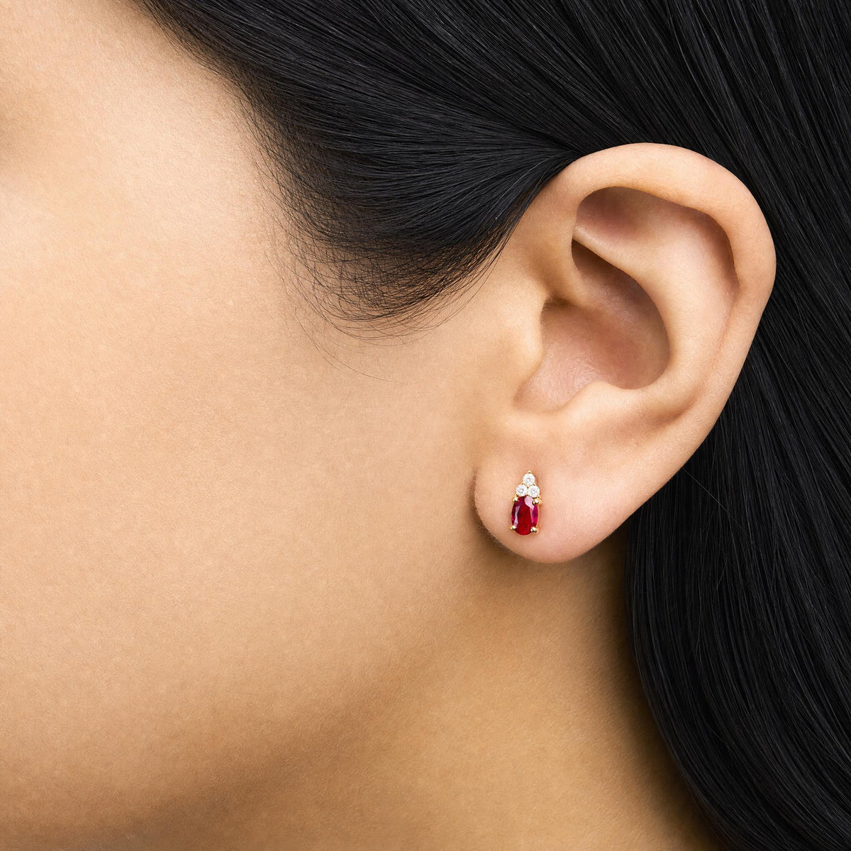 Close-up side view of a model wearing yellow metal earrings featuring oval red gemstones with prong settings, accented by round white gemstones with prong settings above.