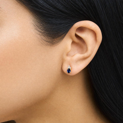 Close-up side view of a model wearing yellow metal earrings featuring an oval blue gemstone set below round white gemstones in a prong setting.