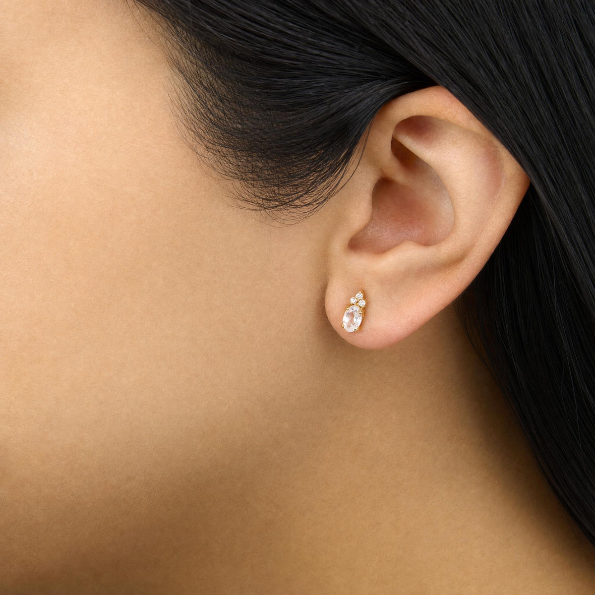 Close-up side view of a model wearing yellow metal earrings featuring an oval-shaped white gemstone with six small round white gemstones clustered above it in prong settings.