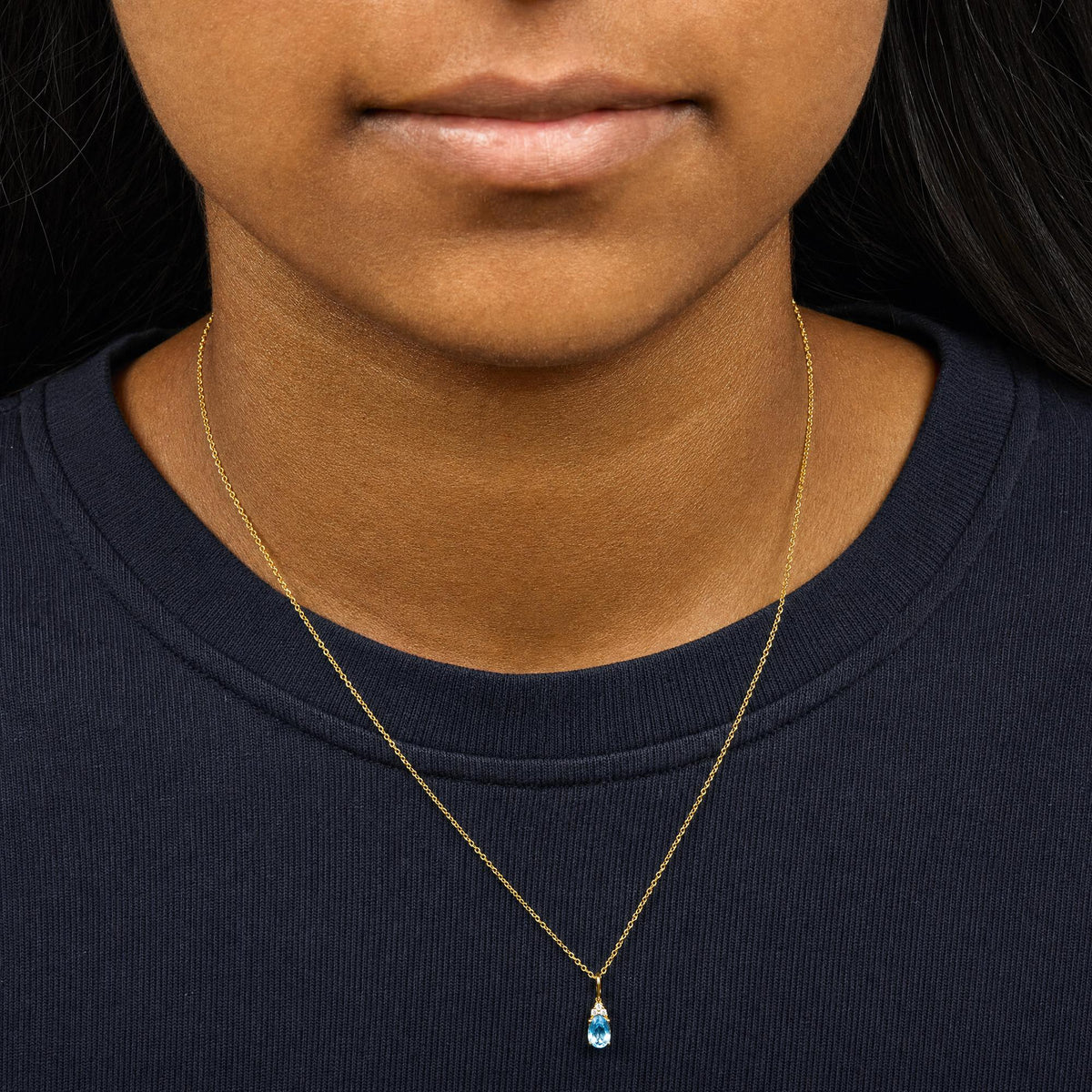 Close-up front view of a model wearing a yellow metal necklace with a pendant featuring a blue oval gemstone and three small white round gemstones set above it.