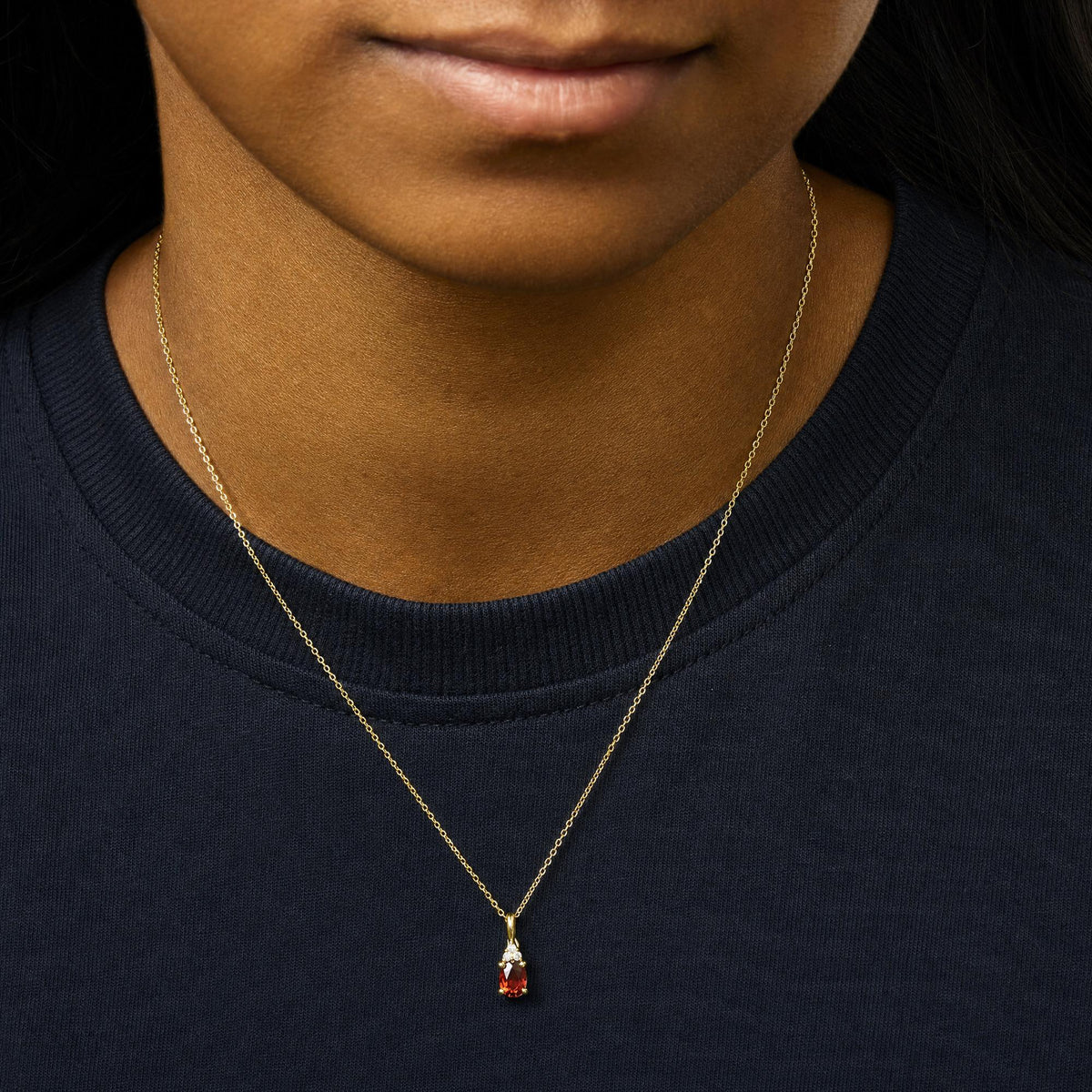 Close-up front view of a necklace with a yellow metal chain featuring a pendant with an oval red gemstone and three round white gemstones above it, shown worn on a model.