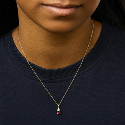 Close-up front view of a necklace with a yellow metal chain featuring a pendant with an oval red gemstone and three round white gemstones above it, shown worn on a model.