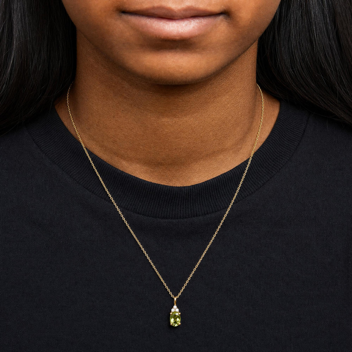 Close-up front view of a model wearing a yellow metal necklace with a pendant featuring an oval green gemstone and three round white gemstones above it.
