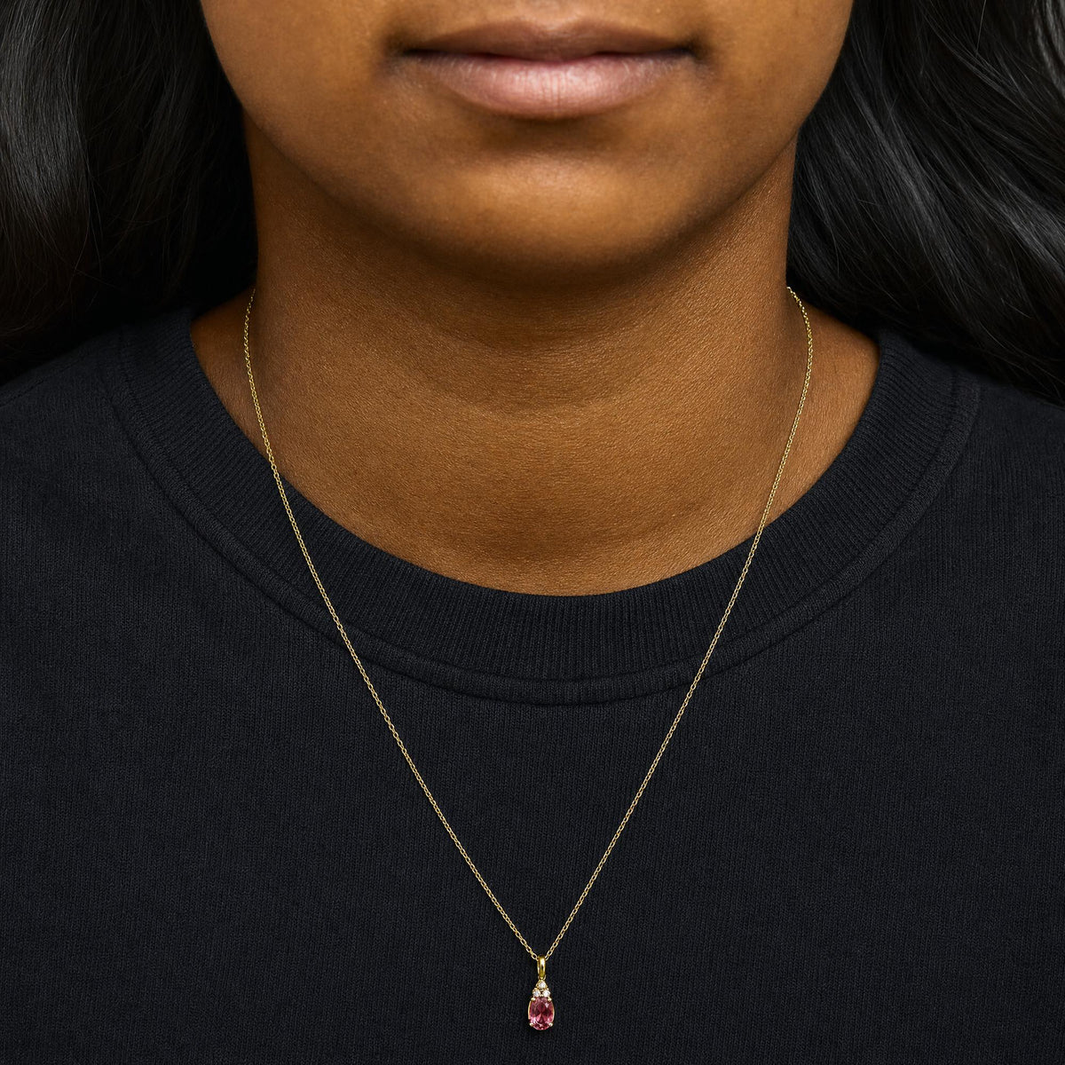 Close-up front view of a model wearing a yellow metal necklace with an oval pink gemstone pendant accented by three small round white gemstones above it.