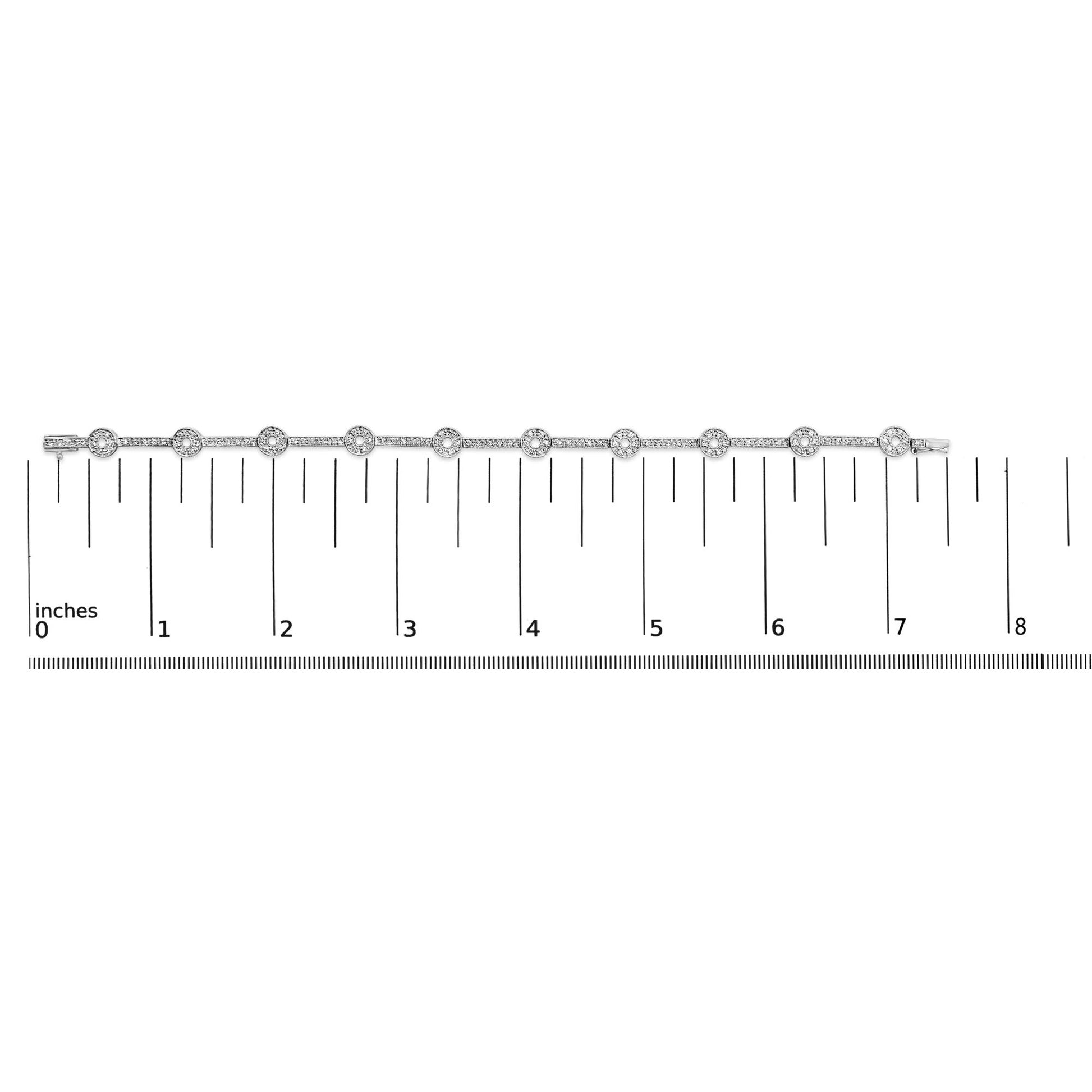 Top-down view of a white metal bracelet with round white gemstones in prong settings, displayed on a ruler for size reference.