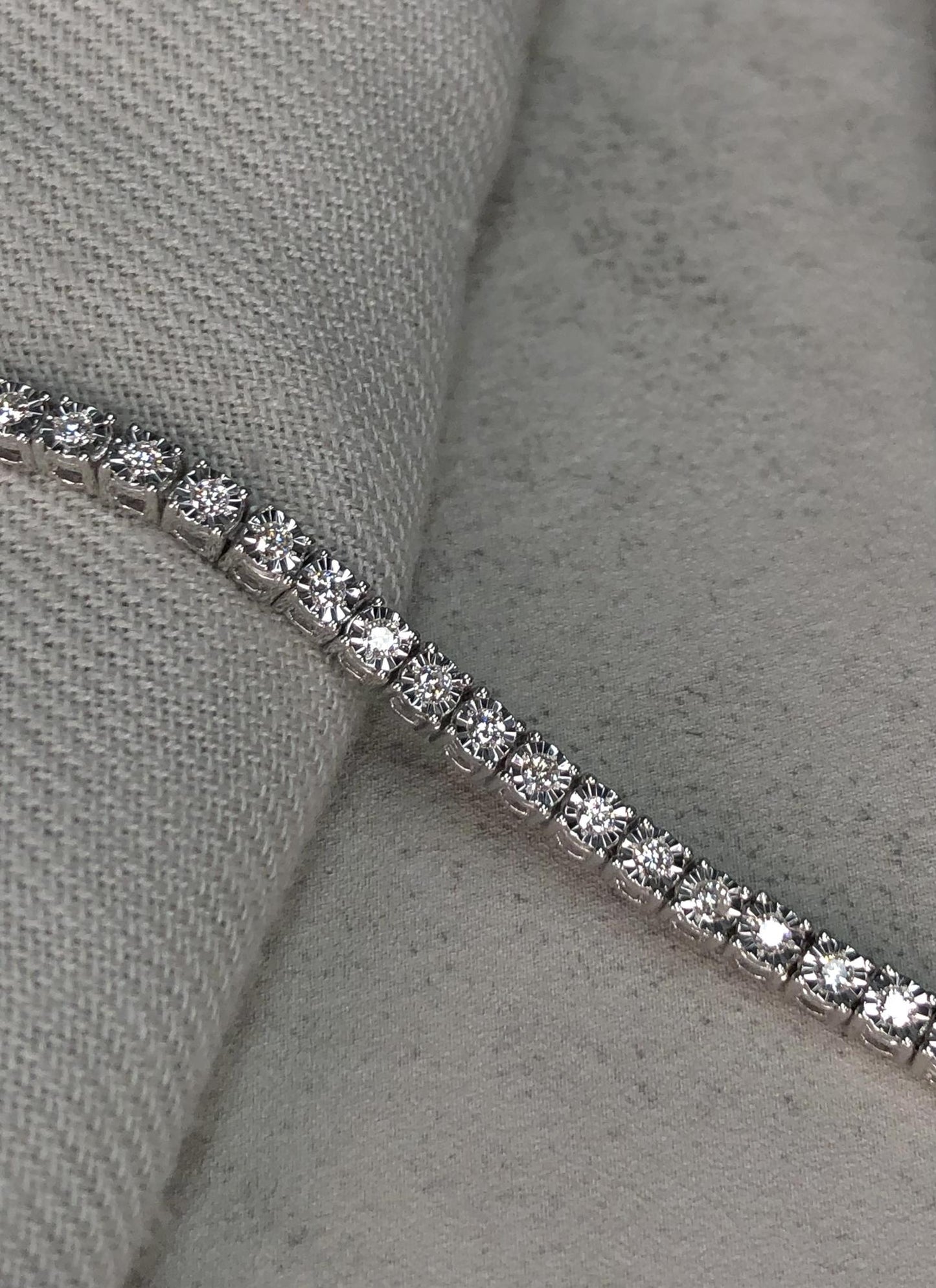 Close-up side angle of a white metal bracelet with round white gemstones in miracle settings, placed on a textured fabric surface.