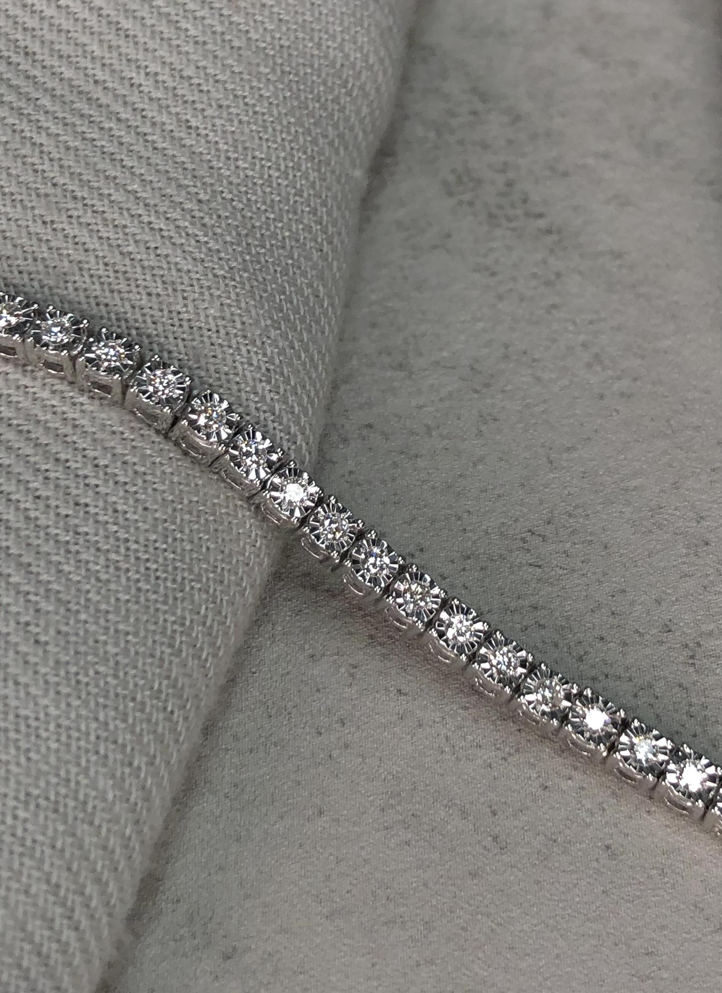 Close-up side angle of a white metal bracelet with round white gemstones in miracle settings, placed on a textured fabric surface.