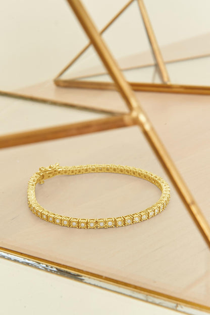 Yellow metal bracelet with round white gemstones in a miracle setting, shown flat on a wooden surface with golden mirrored geometric frames in the background, photographed from a high angle.