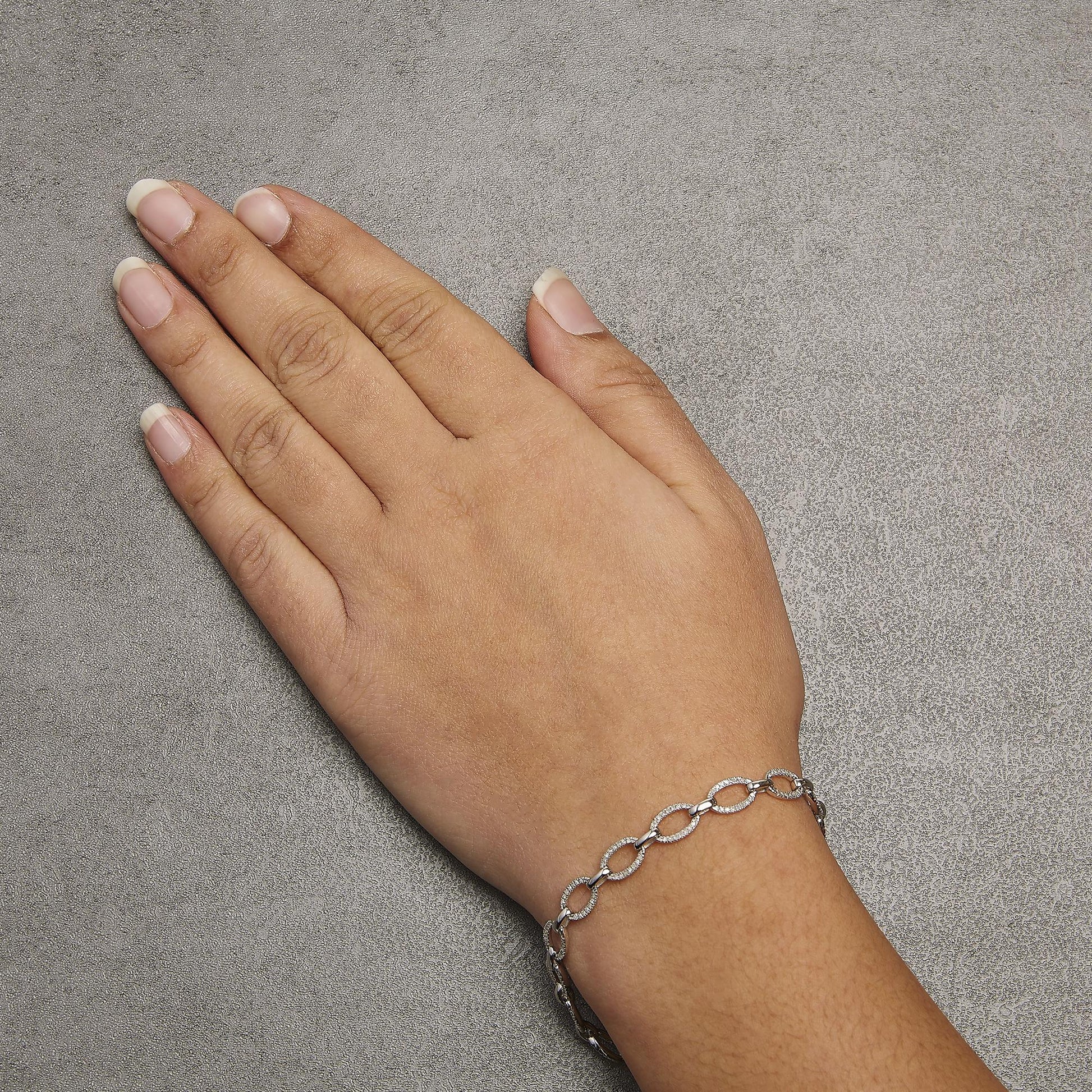 White metal bracelet with oval links encrusted with white gemstones in prong settings, shown on a model's wrist from a top-down angle.